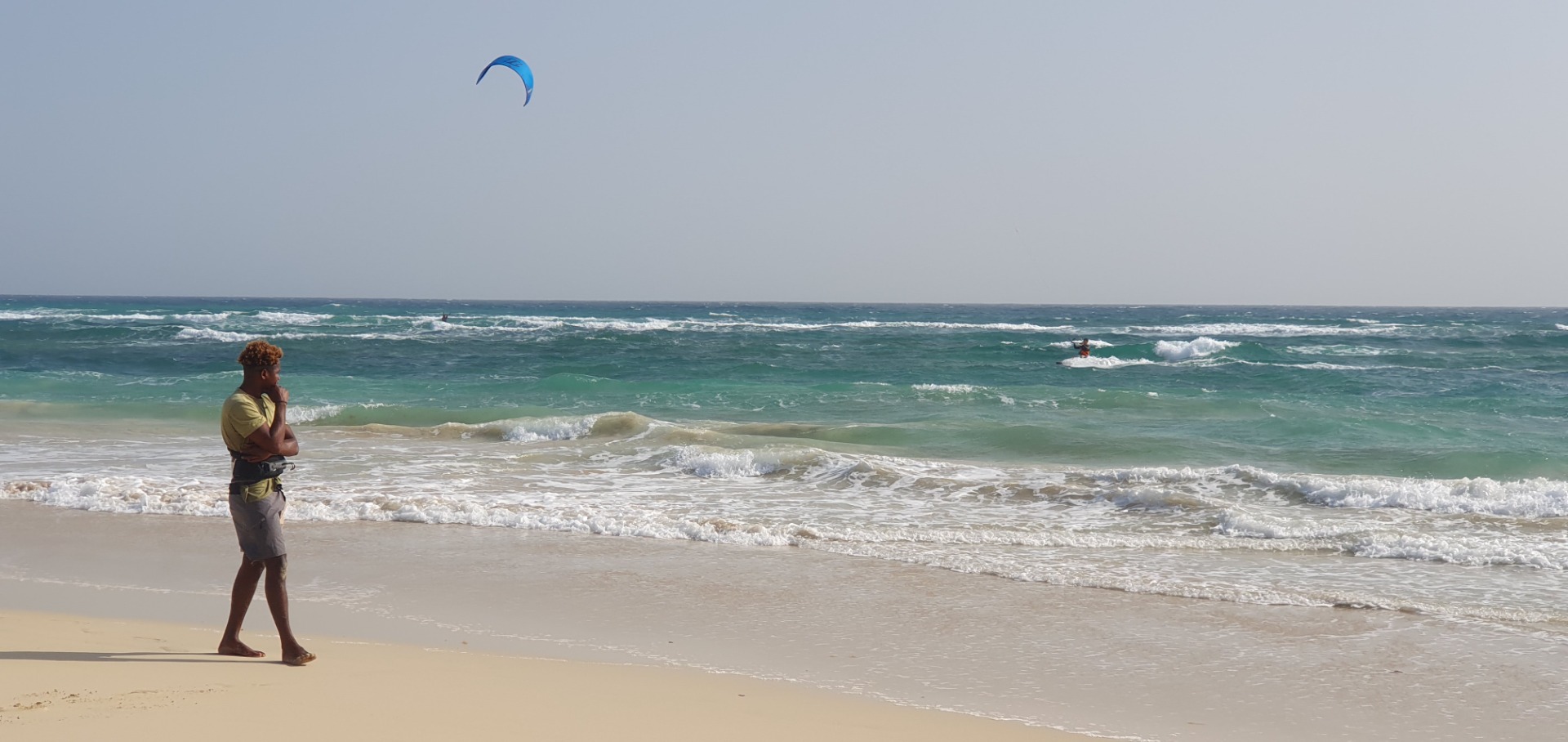 Sal - Cabo Verde - Mann steht am Strand und schaut aufs Meer, während ein Surfer auf einer Welle reitet.