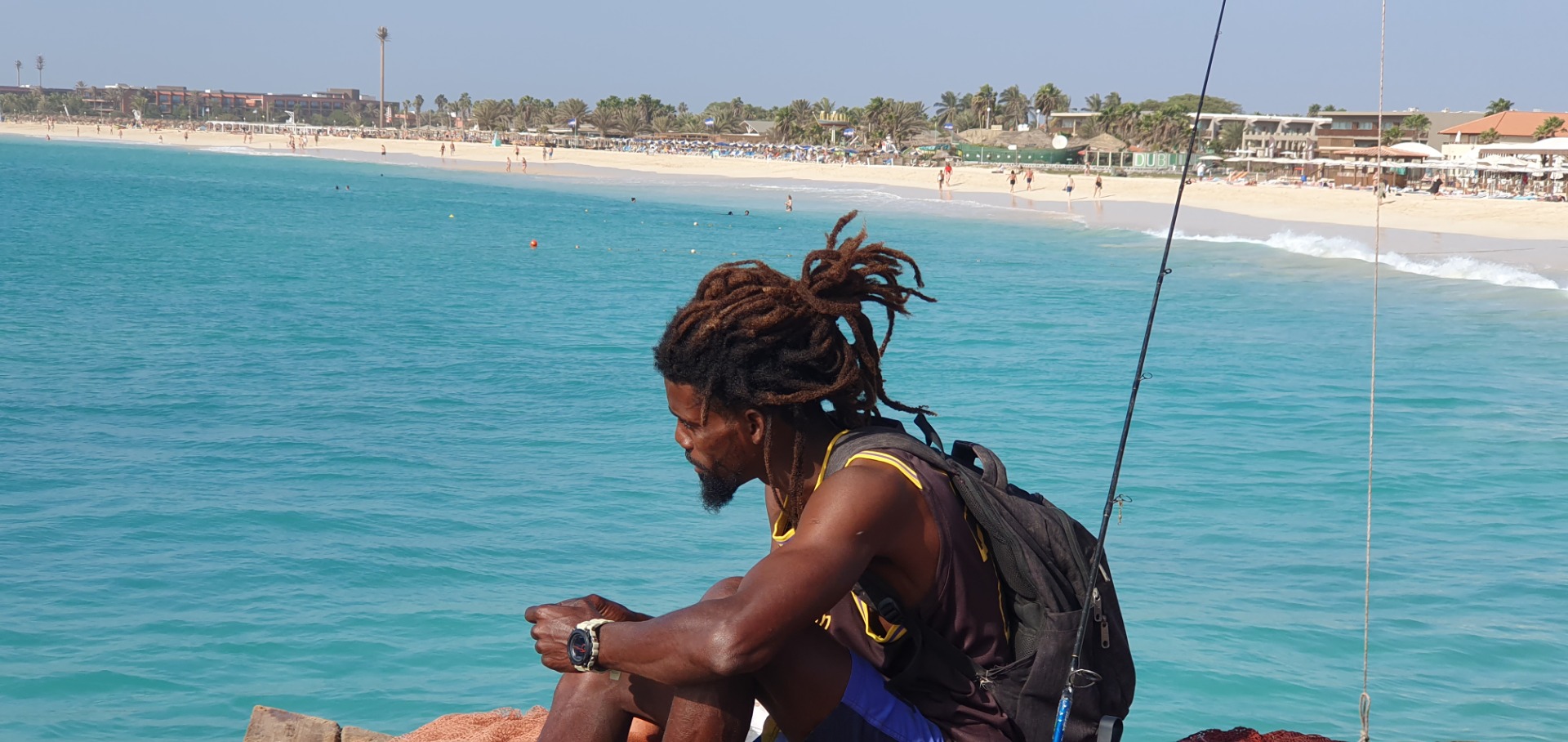 Santa Maria  - Sal - Mann mit Dreadlocks und Rucksack sitzt am Strand, blickt aufs türkisfarbene Meer und die Küste.