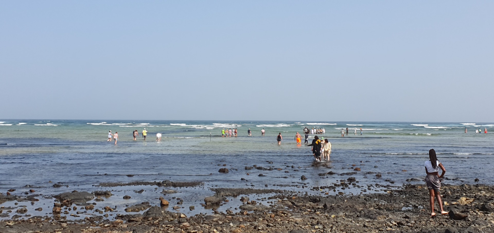 Shark Bay - Sal - Menschen stehen am Strand im flachen Wasser, Wellen rollen über den Küstenbereich, bewölkter Himmel.