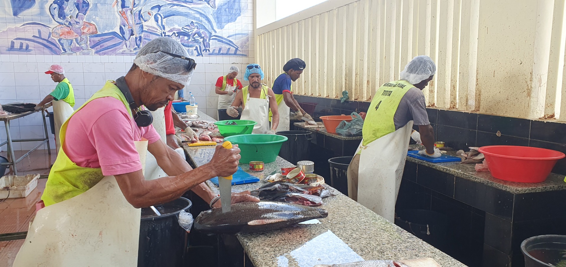 Fischverarbeitungsraum: mehrere Arbeiter mit Schürzen und Hauben reinigen und schneiden Fische an einer Arbeitsbank.
