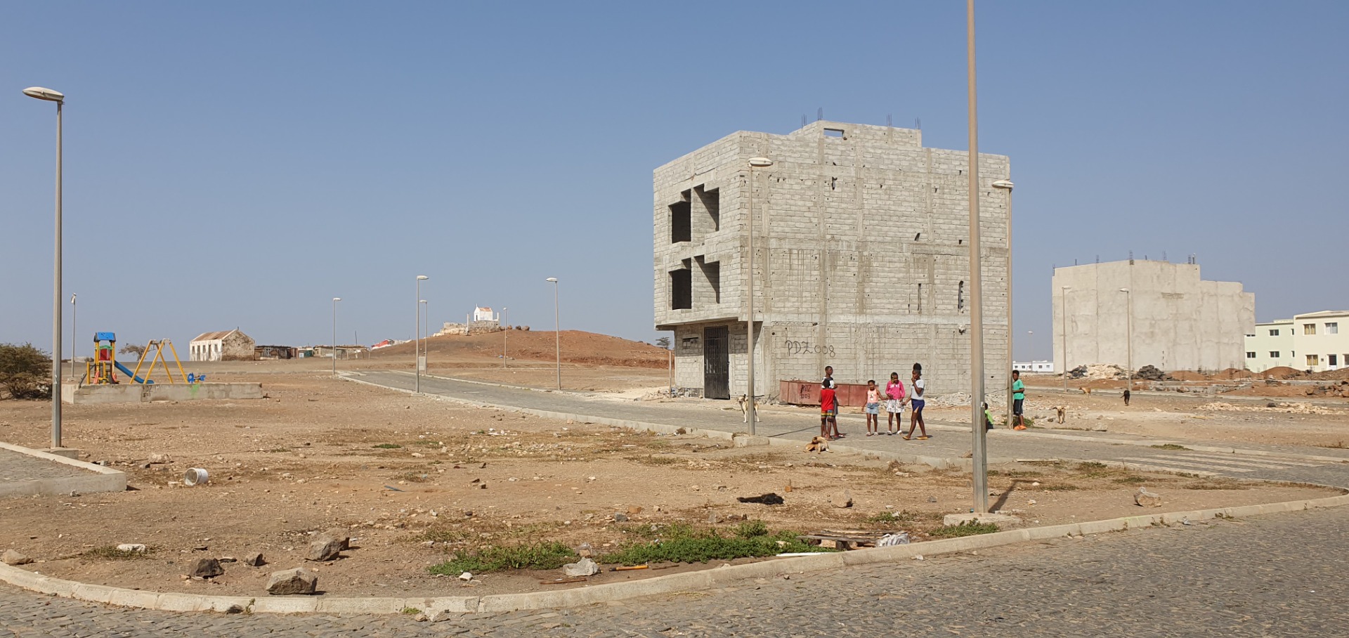 Sal - Cabo Verde -Unfertiges Betonhaus mit offenen Fenstern, Kinder stehen auf Straße, Baustelle in trockener Umgebung.