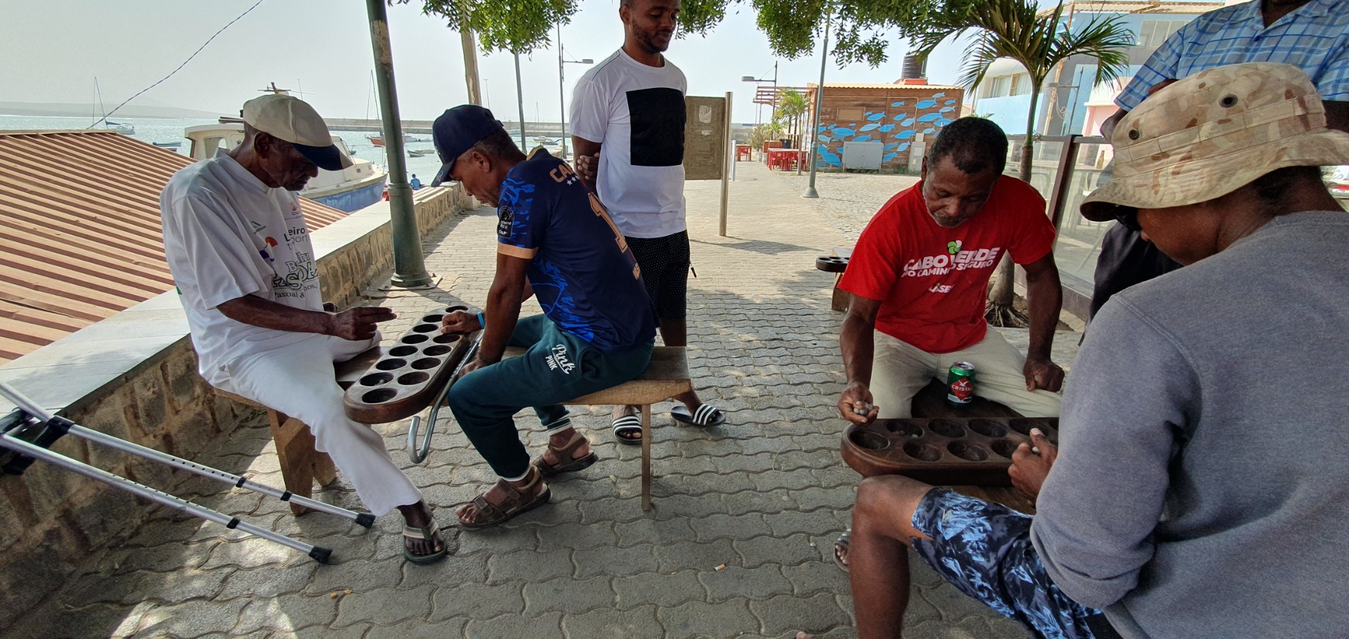 Palmeira - Sal - Männer sitzen am Straßenrand und spielen mit einem Brettspiel; Meer und Gebäude im Hintergrund.