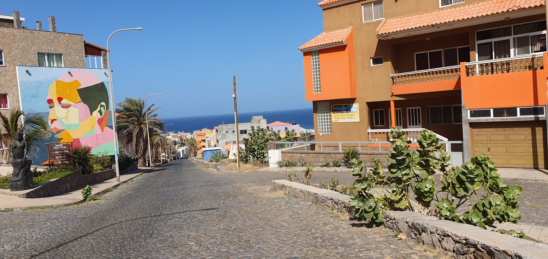 Pontas do Sol - Santo Antao -Straße mit Kopfsteinpflaster, bunten Häusern und Meerblick; Wandgemälde links, Pflanzen und Laternen im Vordergrund.