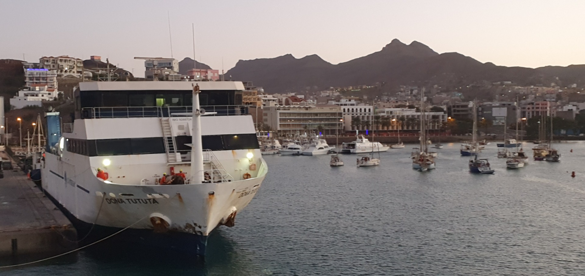 MIndelo - Cabo Verde -Schiff im Hafen vor Küstenstadt, im Hintergrund Berge und zahlreiche Boote bei Dämmerungslicht.