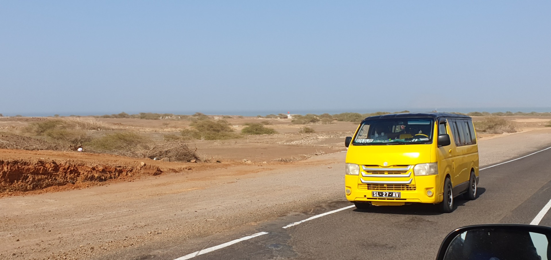 Aluguer Sammeltaxi in Kapverden - Gelber Kleinbus fährt auf einer Straße durch trockene Dünenlandschaft, im Hintergrund blauer Himmel und Sand.